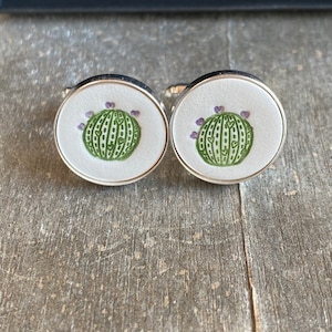 May include: A pair of silver cufflinks with a white background and a green cactus design. The cactus has purple flowers on top.
