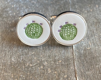 May include: A pair of silver cufflinks with a white background and a green cactus design. The cactus has purple flowers on top.