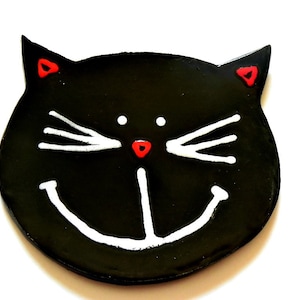 May include: Black ceramic cat face with white whiskers and a white smiling mouth. The cat has red eyes.