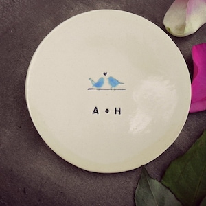 Puede incluir: Un plato de cer&aacute;mica blanco con dos p&aacute;jaros azules que se miran y un peque&ntilde;o coraz&oacute;n encima. El plato tiene las iniciales "A + H" escritas debajo de los p&aacute;jaros.