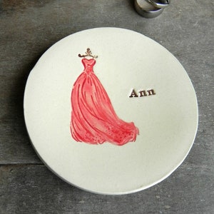 May include: A white ceramic ring dish with a red dress illustration and the name "Ann" written in black.