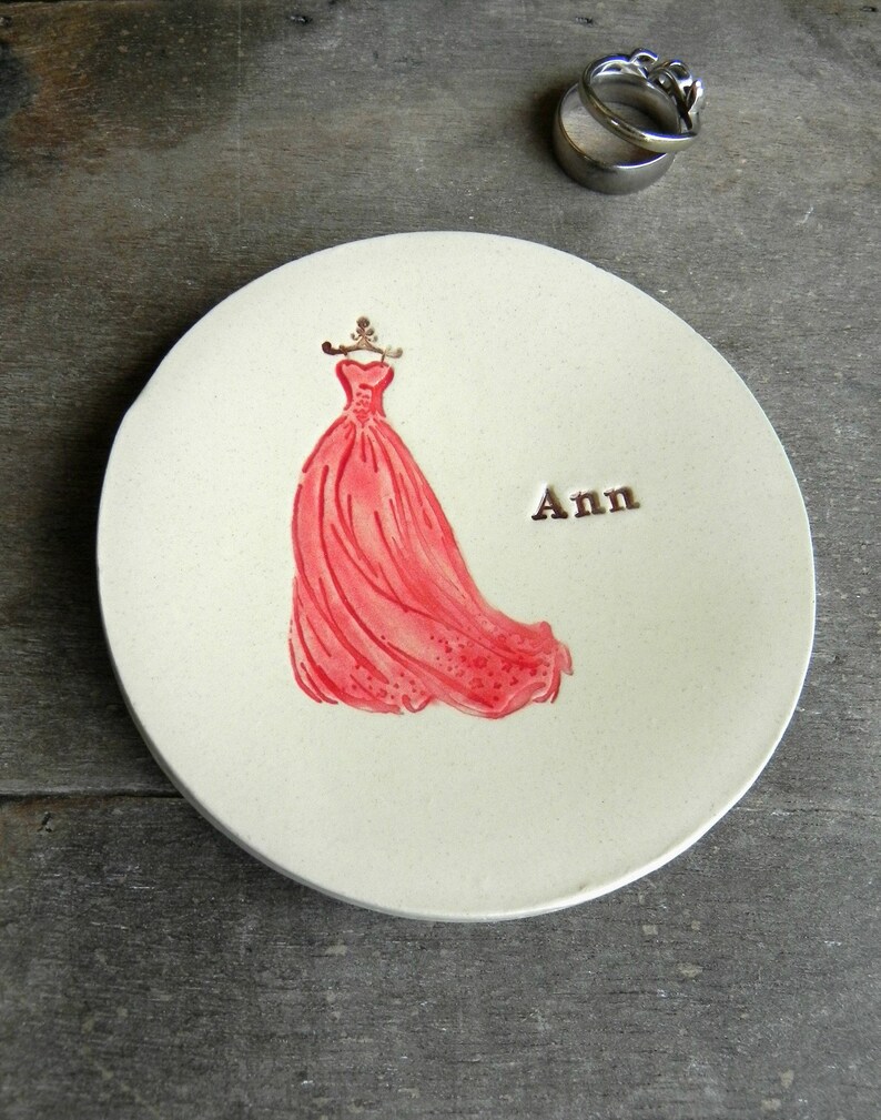May include: A white ceramic ring dish with a red dress illustration and the name "Ann" written in black.