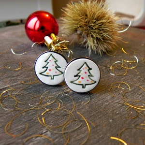 May include: A pair of white ceramic Christmas tree earrings with green outlines and red and yellow polka dots. The earrings are on a wooden surface with gold glitter.