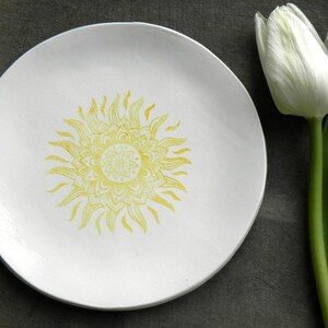 May include: A white ceramic plate with a yellow sun mandala design. The plate is on a gray surface next to a white tulip.