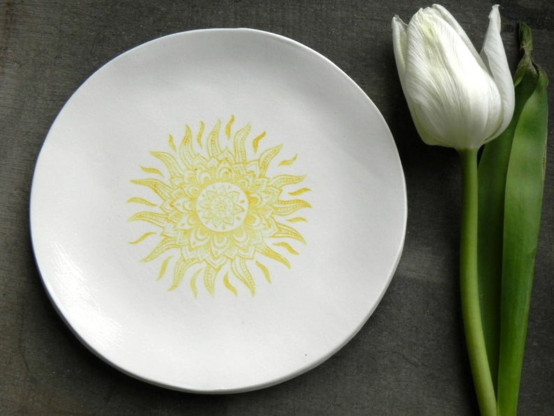 May include: A white ceramic plate with a yellow sun mandala design. The plate is on a gray surface next to a white tulip.