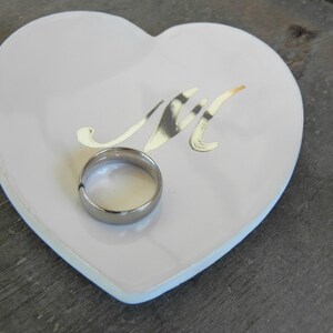 May include: A white heart-shaped ceramic dish with a gold 'M' design and a silver ring resting inside.