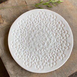 Pode incluir: Prato de cer&acirc;mica branco com um design de renda em relevo. O prato &eacute; redondo e tem um di&acirc;metro de aproximadamente 23 cm.