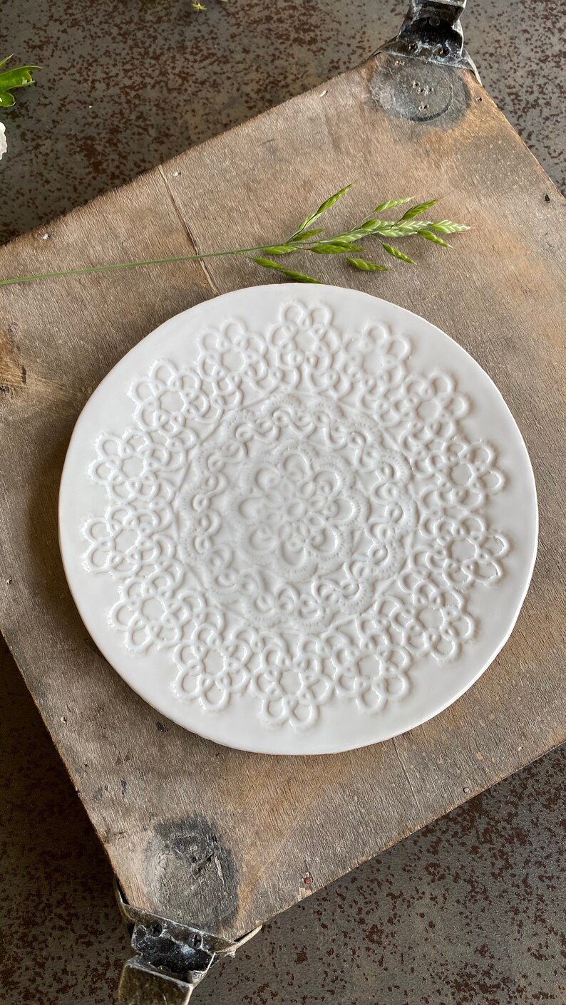 Pode incluir: Prato de cer&acirc;mica branco com um design de renda em relevo. O prato &eacute; redondo e tem um di&acirc;metro de aproximadamente 23 cm.