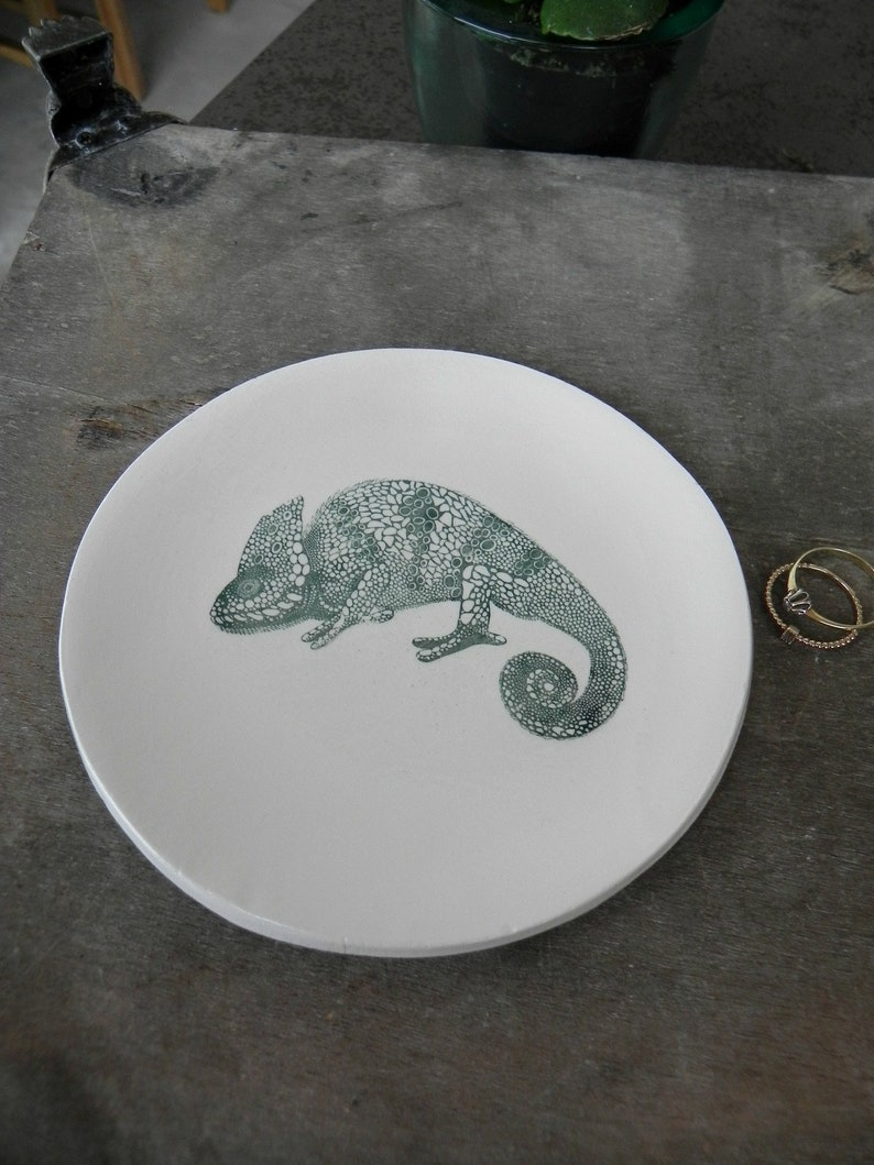 Puede incluir: Un plato de cer&aacute;mica blanco con un dise&ntilde;o de camale&oacute;n verde. El plato es redondo y tiene un borde ligeramente elevado.