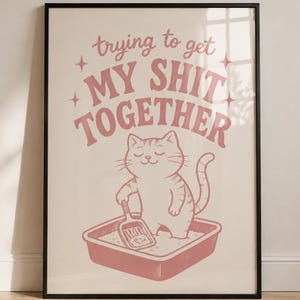 Könnte beinhalten: Ein gerahmter Kunstdruck mit einer hellrosa Illustration einer Katze, die in einer Katzentoilette steht und eine Schaufel hält. Der Text "trying to get MY SHIT TOGETHER" ist in Rosa über der Katze gedruckt und mit vier kleinen Sternen verziert.