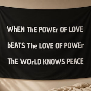 Könnte beinhalten: Ein rechteckiger schwarzer Wandbehang mit weißem Text: "WhEN ThE POWEr of LOVE bEATS The LOVE OF POWEr The WOrLD KNOWS PEACE". Der Stoff ist mit Metallösen an den vier Ecken an einer neutralen Wand befestigt.