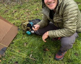Può includere: Una persona che indossa un piumino con cappuccio verde oliva e pantaloni grigio scuro, tiene in mano un fascio di picchetti neri con punte blu brillante, inginocchiata accanto a una tenda marrone con tiranti gialli fissati sull'erba verde.