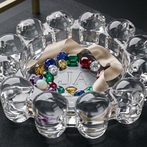 May include: A clear glass bowl with a scalloped, bubble-like rim, featuring the engraved letters JA at the center. The bowl contains a collection of multi-colored faceted gemstones in shades of red, green, blue, and yellow, accented by a beige ribbon.
