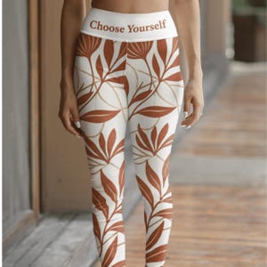 May include: High-waisted leggings featuring a white base with a repeating rust-colored botanical leaf pattern. The wide waistband displays the text Choose Yourself in a brown font. These fitted leggings extend to the ankles with a consistent print.