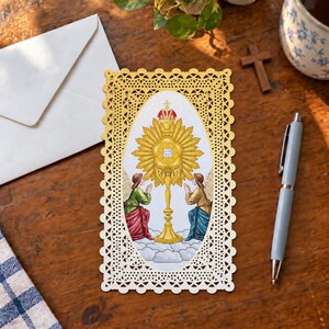 Puede incluir: Una tarjeta de oración rectangular con un borde decorativo de encaje troquelado en color dorado. El centro presenta una ilustración ovalada de una custodia dorada con una cruz, flanqueada por dos ángeles arrodillados con túnicas sobre nubes.