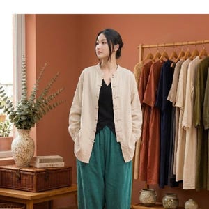 Può includere: Una camicia beige a maniche lunghe con colletto alla coreana e chiusura frontale con bottoni in tessuto. La camicia è indossata sopra un top nero senza maniche e pantaloni ampi color verde acqua, creando un outfit a strati dai toni neutri.
