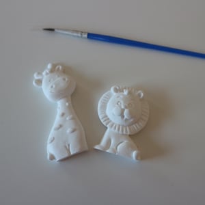 Peut inclure: Deux figurines d'animaux en céramique blanche non peinte, l'une en forme de girafe et l'autre en forme de lion, placées à côté d'un pinceau bleu fin avec une virole argentée. Les figurines présentent des détails en relief du visage et de la crinière.