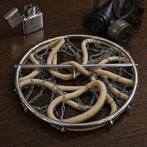 Puede incluir: Un marco circular de metal que contiene una cuerda beige enrollada y cadenas de metal plateado, acompañado de un encendedor de metal plateado y una máscara de gas de goma negra con un filtro verde, dispuestos sobre una superficie oscura.