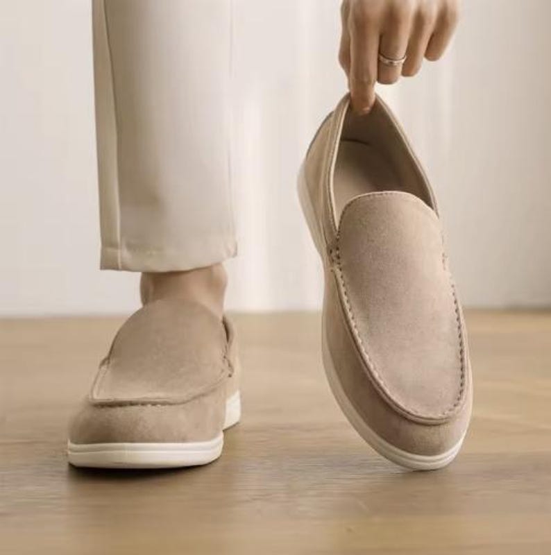 May include: A pair of beige slip-on loafers featuring a soft suede-like texture, a rounded toe, and a thick white rubber sole. The shoes have visible tonal stitching along the vamp and sides, providing a clean, neutral look for casual footwear.