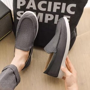 May include: A pair of dark gray slip-on loafers featuring a suede-like texture, a rounded toe, and a thick white rubber sole. The shoes have visible stitching along the top vamp and a simple, low-profile design with a light-colored interior lining.