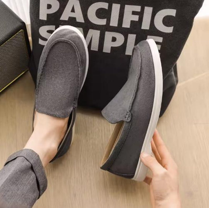 May include: A pair of dark gray slip-on loafers featuring a suede-like texture, a rounded toe, and a thick white rubber sole. The shoes have visible stitching along the top vamp and a simple, low-profile design with a light-colored interior lining.