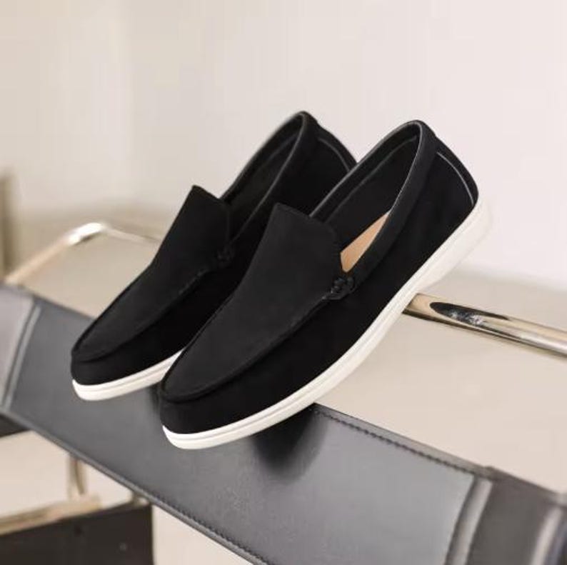 May include: A pair of black slip-on loafers featuring a smooth matte finish, a rounded toe design, and a thick white rubber sole. The shoes have a clean, low-profile silhouette with visible stitching along the vamp and a simple, unadorned exterior.