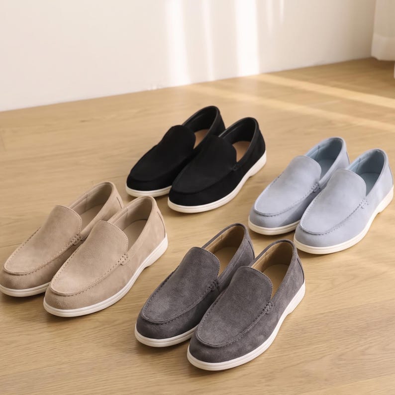 May include: Four pairs of slip-on loafers in black, light blue, beige, and dark gray. Each pair features a suede-like texture, a rounded toe, visible stitching along the vamp, and a solid white rubber sole. The shoes are arranged in a group.