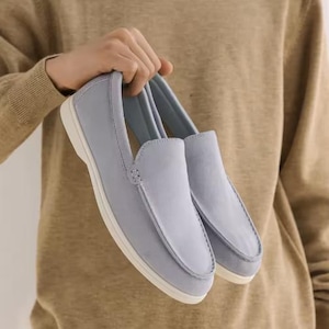 May include: A pair of light blue slip-on loafers featuring a smooth matte finish, a rounded toe box with visible tonal stitching along the vamp, and a thick white rubber sole. The shoes have a low-cut collar and a simple, streamlined design.