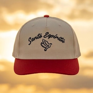 May include: A beige baseball cap with a red brim and a red top button. The front panel features black embroidered text that reads Santo Espíritu above a black embroidered graphic of a flying dove with wings spread in a downward motion.