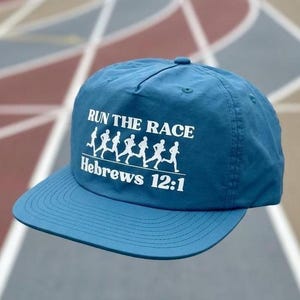 May include: A blue baseball cap with a flat brim featuring white screen-printed text that reads RUN THE RACE and Hebrews 12:1, accompanied by a graphic of six silhouettes of people running in a line across the front panel of the hat.
