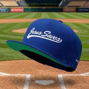 May include: A royal blue baseball cap featuring the white embroidered text Jesus Saves in a script font across the front crown. The cap has a flat, bright green brim and a structured five-panel design with a small white logo on the side panel.