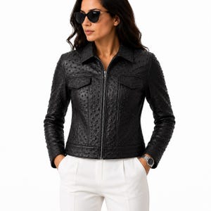 May include: A black jacket with a textured, dotted surface pattern, featuring a collared neckline, a full-length front zip, and two chest pockets with flaps. The jacket has long sleeves and a structured fit, worn with plain white trousers.