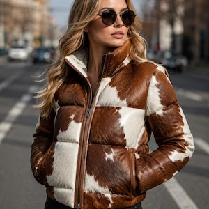 Könnte beinhalten: Eine Steppjacke mit braun-weißem Kuhfellmuster, hohem Kragen und durchgehendem Frontreißverschluss. Die Jacke hat eine strukturierte Oberfläche und wird mit einer dunklen Sonnenbrille getragen. Das Design umfasst horizontal gesteppte Abschnitte.