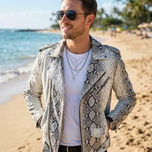 May include: A white and grey snakeskin-patterned leather motorcycle jacket with silver-tone zips, a notched lapel, and a belted waist, worn over a plain white t-shirt with layered silver chain necklaces and dark blue denim jeans.