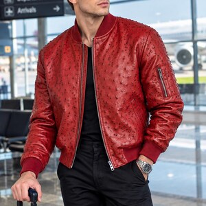 Peut inclure: Un blouson aviateur en cuir rouge texturé avec un col, des poignets et un ourlet côtelés. Le vêtement présente une fermeture éclair frontale, une poche utilitaire zippée sur la manche gauche et une texture de surface à motifs de plumes.