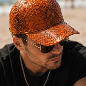 Puede incluir: Una gorra de béisbol con un patrón de piel de serpiente texturizado en color naranja-marrón. La gorra tiene una corona estructurada, una visera curva y un pequeño botón en la parte superior central. El material muestra una textura detallada.