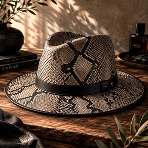 May include: A fedora hat featuring a beige and black snakeskin pattern throughout the crown and wide brim. The hat is accented with a solid black leather band around the base of the crown, which includes a small metallic stud detail on the side.