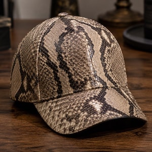 Pode incluir: Um boné de beisebol com um padrão texturizado de pele de cobra em tons de bege, castanho claro e castanho escuro. O boné tem uma copa estruturada, uma aba curva e um pequeno botão no centro do topo. O material apresenta um aspeto de escamas.