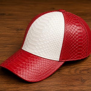 Puede incluir: Una gorra de béisbol con un patrón texturizado similar a escamas. El panel frontal es blanco, mientras que los paneles laterales y la visera curva son de un rojo vibrante. El diseño incluye un botón en la parte superior y costuras.