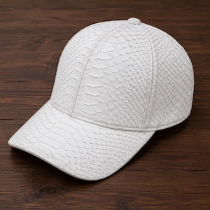 May include: A white baseball cap featuring a textured snakeskin pattern across the entire surface. The cap has a structured crown, a curved brim, and a small button at the top center. The material displays a detailed scale-like finish.
