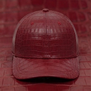 Peut inclure: Une casquette de baseball rouge foncé présentant un motif texturé en relief façon peau de crocodile sur toute la surface. La casquette possède une calotte structurée, une visière incurvée et un petit logo discret en relief sur le panneau avant.