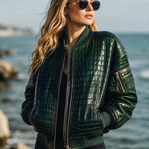 May include: A dark green bomber jacket featuring a glossy, embossed crocodile skin texture. The jacket includes a front zipper closure, ribbed knit collar and cuffs, and a small zippered utility pocket on the upper left sleeve with gold hardware.