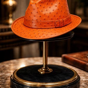May include: A vibrant orange fedora hat featuring a textured ostrich leather pattern with a matching orange band and a small rectangular buckle detail. The hat has a structured crown and a wide brim, showcasing a distinct dotted surface texture.