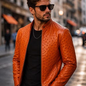 Può includere: Una giacca in pelle color arancione vivace con una superficie testurizzata a pois distintivi. La giacca presenta un colletto alla coreana, una cerniera frontale a tutta lunghezza e maniche lunghe. Indossata sopra una t-shirt nera con una catenina.