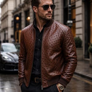 May include: A brown textured leather bomber jacket with a ribbed collar and cuffs, worn over a black button-down shirt. The jacket features a distinct dotted pattern across the surface, a front zipper closure, and is paired with black trousers.