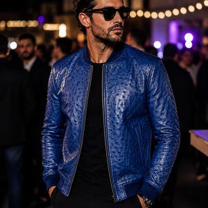May include: A vibrant blue bomber jacket featuring a textured, quill-patterned leather finish, a central silver zipper, and dark blue ribbed cuffs and collar. The jacket is worn over a plain black t-shirt and paired with black trousers.