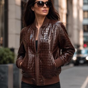 May include: A dark brown bomber jacket featuring a glossy, embossed crocodile skin texture. The jacket includes a front zipper closure, ribbed brown cuffs, a ribbed hem, and two side pockets. The garment is worn over a simple black top.