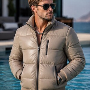 May include: A beige puffer jacket featuring a high collar, a full-length black front zip, and a single vertical zipped chest pocket. The jacket includes two side pockets with black zips, ribbed cuffs, and a smooth, quilted exterior finish.