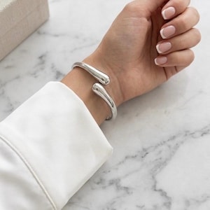 Può includere: Un bracciale rigido in metallo argentato lucido caratterizzato da un design aperto con due estremità arrotondate a forma di goccia. La superficie metallica è liscia e riflettente, creando una forma fluida e continua attorno al polso.