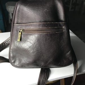 May include: A dark brown leather backpack featuring a front zippered pocket with a gold-toned metal zipper pull embossed with the text BASS. The bag has a rounded top, a smooth texture, and adjustable shoulder straps attached to the base.